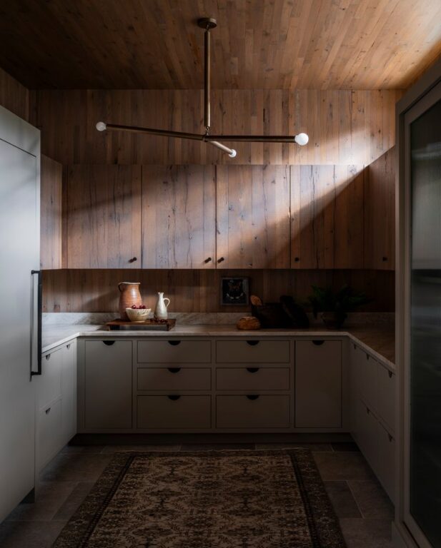 A modern kitchen with a reclaimed wood wall and ceiling, gray cabinets, a patterned rug, and a minimalist light fixture. Sunlight streams in, highlighting a countertop with pottery, bread, and decorative items.