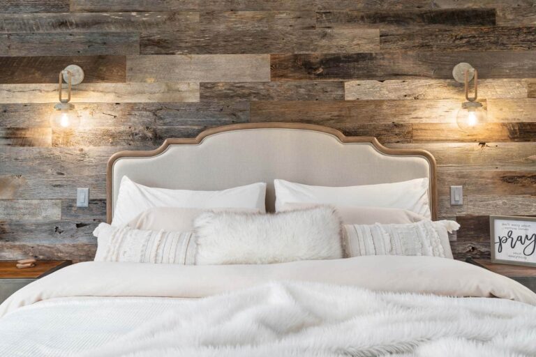 A cozy bed with white bedding and pillows is set against a rustic reclaimed wood wall. Two wall-mounted lamps with exposed bulbs are on each side of the bed, and a small framed sign sits on the right nightstand.