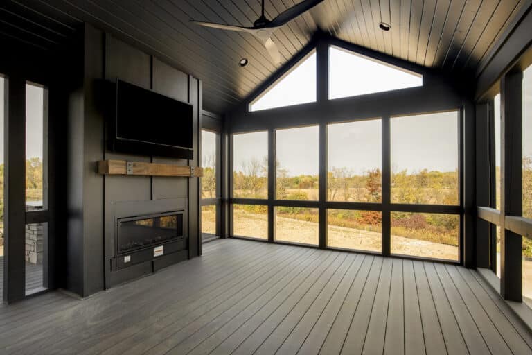 Modern screened-in porch with dark wood floors and ceiling, large windows offering views of trees and fields, a mounted TV above a sleek wood fireplace mantel, and a ceiling fan. The room is empty and filled with natural light.
