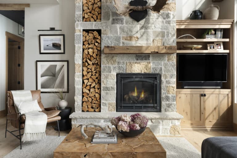 Rustic living room featuring a stone fireplace with a wood fireplace mantel, stacked firewood niches, mounted antlers, built-in shelving with TV, leather armchair, wood slab coffee table, and dried floral arrangement.