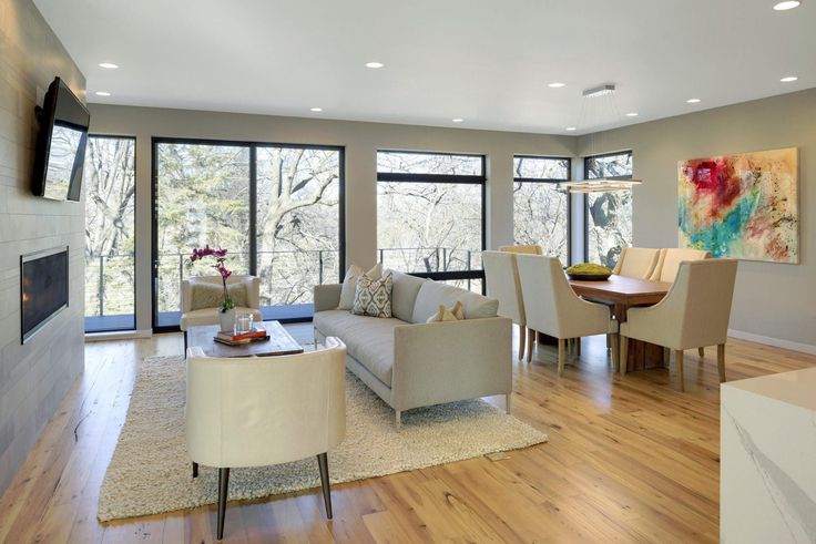Modern open-concept living and dining room with large windows, warm wood floors, cream furniture, a cozy rug, wall-mounted TV, and colorful abstract art on the wall. Trees are visible outside.