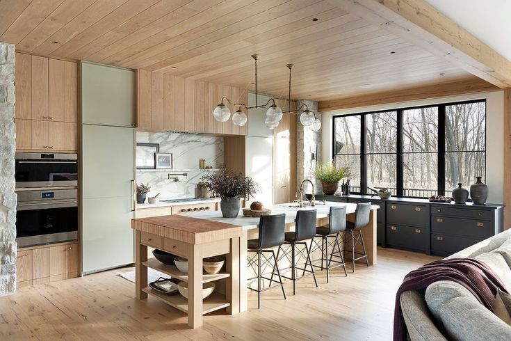 Modern kitchen with light wood cabinets, marble backsplash, wood floors, a large island with barstools, pendant lights, and black window frames overlooking trees. A wooden butcher block and dark lower cabinets add contrast.