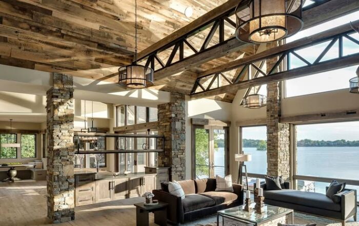 Spacious living room with rustic wood ceiling, exposed beams, stone pillars, antique elm wood floors, large windows, modern furniture, and a view of the lake. Natural light fills the open, airy space decorated in neutral tones.
