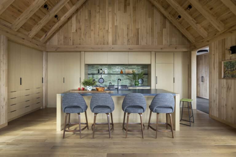An example of biophilic and global design. It's modern kitchen with wooden walls and high vaulted ceiling, blending Japanese design elements, a central island with four gray barstools, green tile backsplash, countertop with plants and kitchen items, and a painting on the wall to the right.