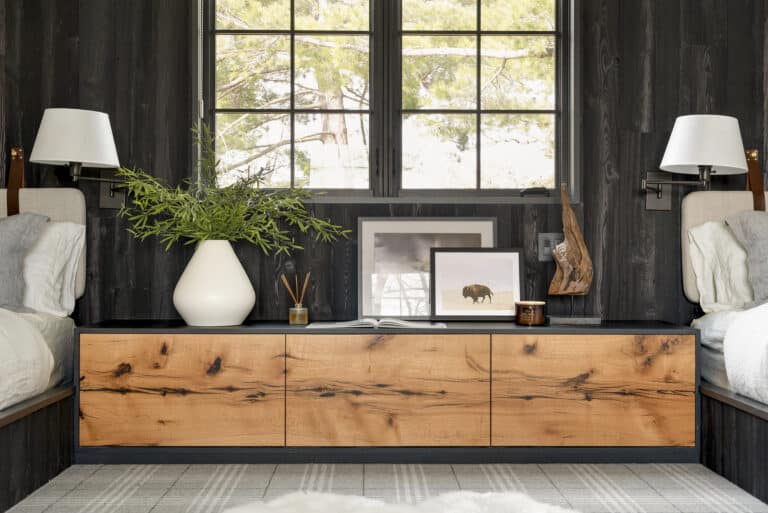 A modern living space that features elements of global design. A black wall with a large window, a wooden cabinet, Japanese decor accents, a white vase with greenery, framed art, and two beds on either side, each with white bedding and wall-mounted lamps.