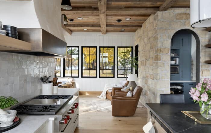 A cozy kitchen with a gas stove and rustic wooden beams opens into a bright living area with leather armchairs, large windows, stone walls, and a view of autumn trees—echoing the charm found in classic European style homes and other global design styles.