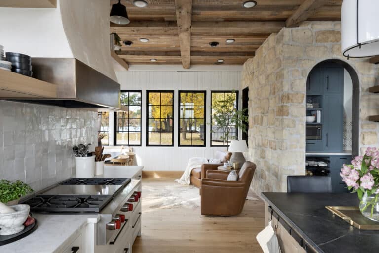 A cozy kitchen with a gas stove and rustic wooden beams opens into a bright living area with leather armchairs, large windows, stone walls, and a view of autumn trees—echoing the charm found in classic European style homes and other global design styles.