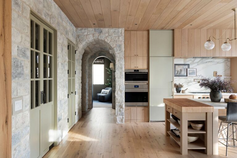 A bright kitchen with white oak wood floors, stone walls, built-in appliances, a marble backsplash, a wooden island with open shelves, and an arched stone doorway leading to a cozy room with a blue armchair.