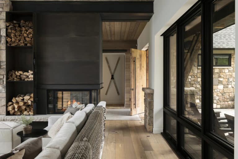 A modern rustic living room with a fireplace, stacked firewood, a cozy sectional sofa, wood floors, open doorway, and large windows overlooking a stone exterior.