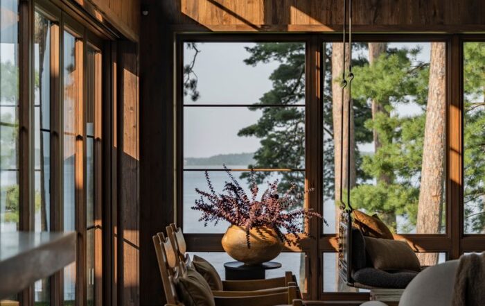 A cozy, modern living room with large windows overlooking a lake and tall pine trees, natural wood walls, comfortable chairs, a gray sofa, and a vase with purple flowers on a central table.