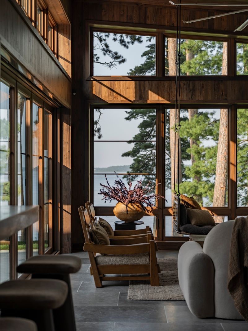 DnEe0_Zw A cozy, modern living room with large windows overlooking a lake and tall pine trees, natural wood walls, comfortable chairs, a gray sofa, and a vase with purple flowers on a central table.