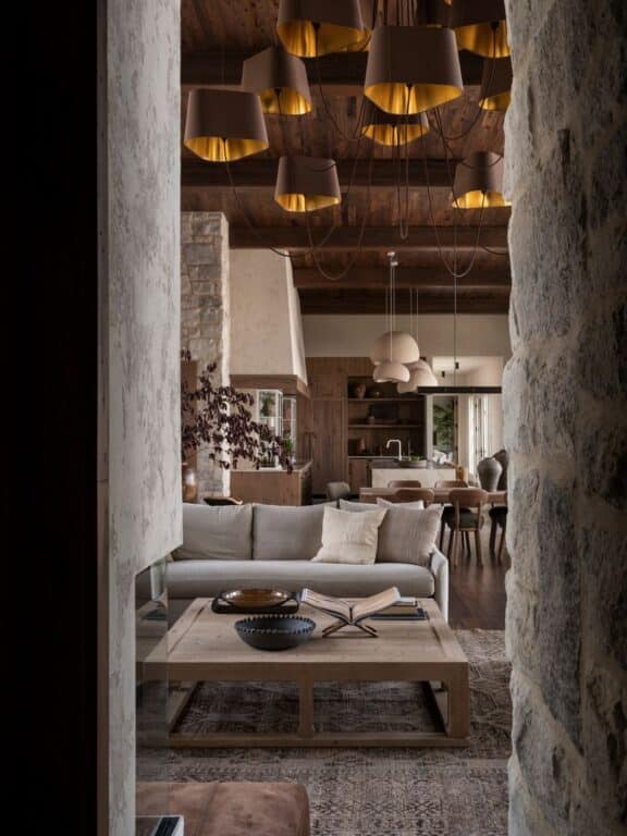 Cozy living room with stone walls, a beige sectional sofa, and a wooden coffee table with books and bowls. Modern pendant lights hang from a wooden ceiling, and the space opens to a dining area and kitchen in warm, earthy tones.