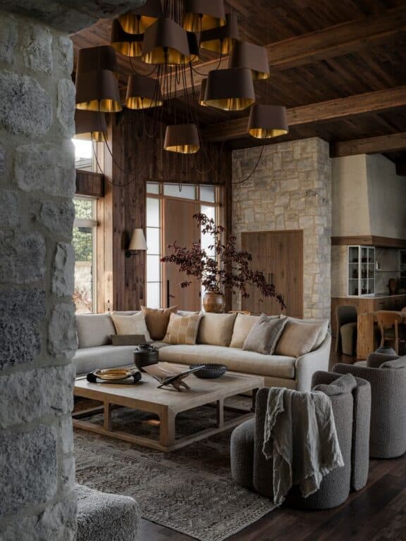 A cozy living room with a large beige sectional sofa, textured armchairs, a wooden coffee table, stone walls, wooden ceiling, and a modern chandelier with brown lampshades. Natural light fills the rustic, elegant space.