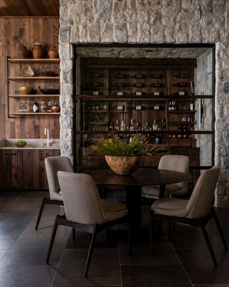 A cozy dining area with four cushioned chairs around a round table, a large wooden bowl with plants as a centerpiece, stone walls, wooden shelves with pottery, and a wall displaying wine bottles in the background.