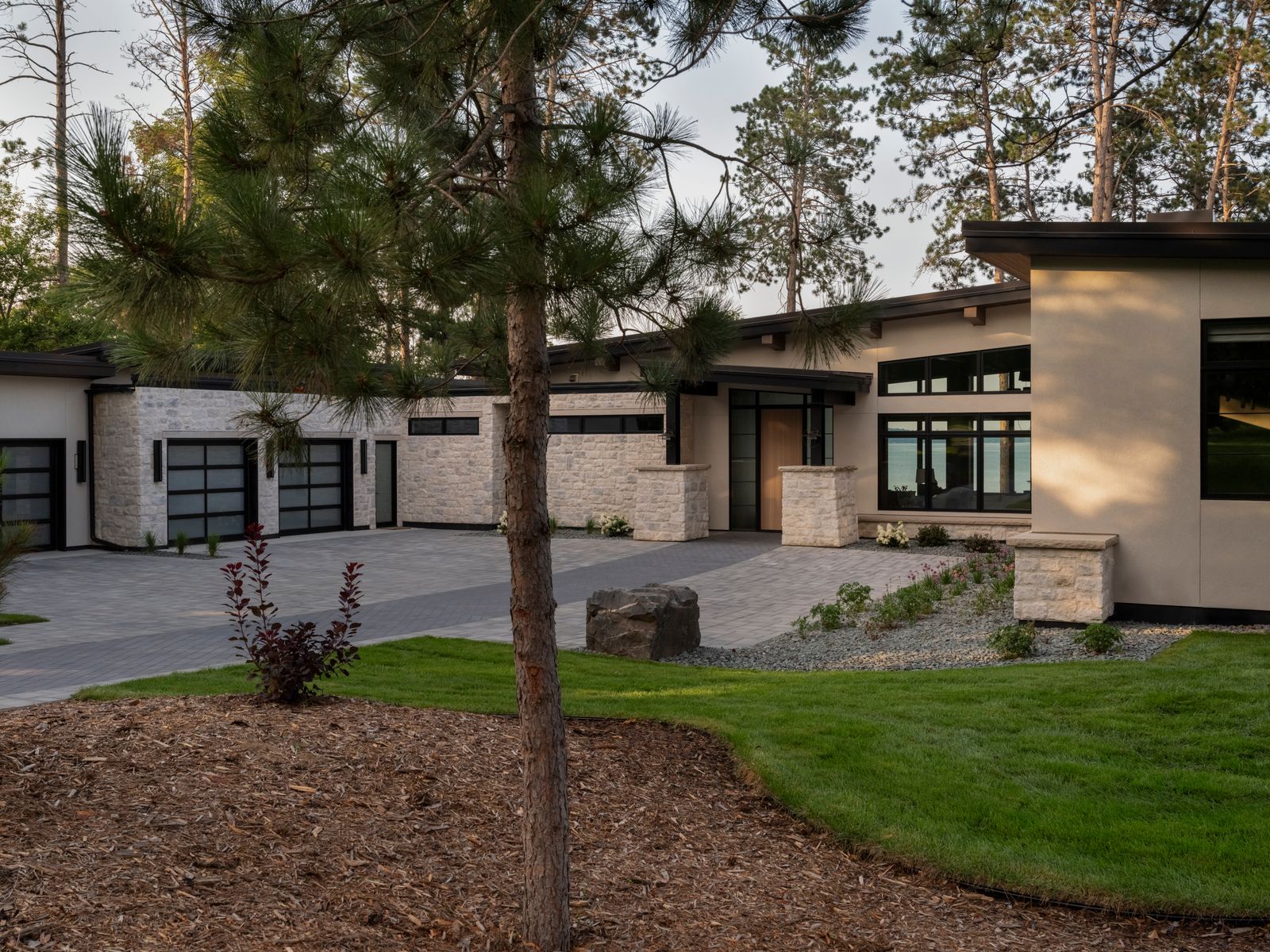 Modern single-story house with large windows, stone and stucco exterior, and a spacious driveway. The yard features neatly landscaped grass, mulch, small plants, and a pine tree in the foreground. Tall trees are visible in the background.
