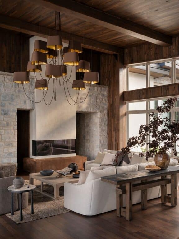 A cozy living room with rustic wood walls and ceiling, stone fireplace, large windows, neutral sofas, a cluster of brown pendant lights, and a wooden coffee table with decor items and a plant in a vase.