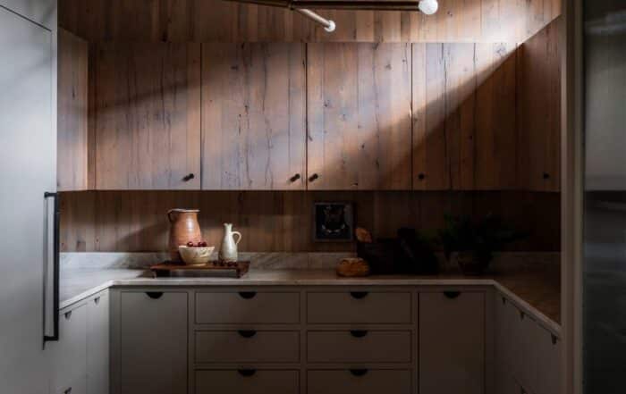 A modern kitchen with wood-paneled walls and ceiling, light gray cabinets, a patterned rug, minimalist lighting, and a marble countertop with decorative items and kitchenware arranged neatly.