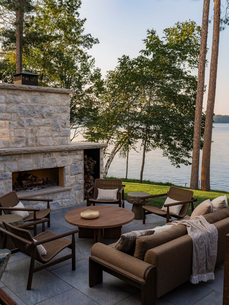 Outdoor patio with a stone fireplace, brown cushioned chairs and sofa, round wooden table, surrounded by trees overlooking a calm lake at sunset. A cozy blanket is draped over the sofa.
