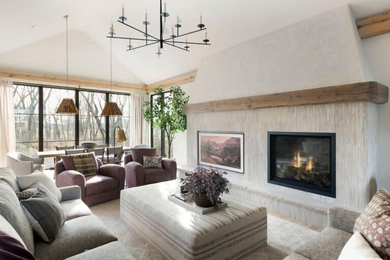 A bright, modern living room with a fireplace, neutral sofas, armchairs, a striped ottoman, large windows with natural light, a dining area in the background, and a potted plant beside the dining table.