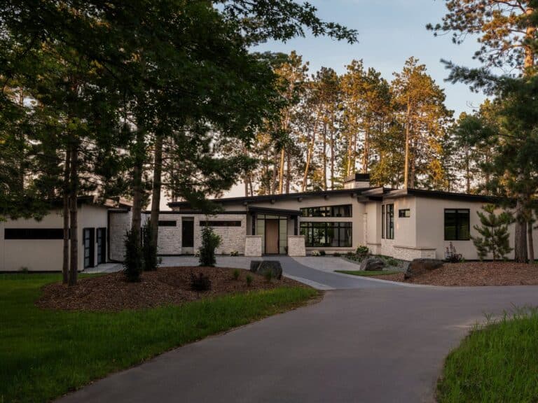 A modern, single-story house with large windows, light-colored stone siding, and a flat roof, surrounded by tall trees and landscaped with mulch and grass, seen from a paved driveway.