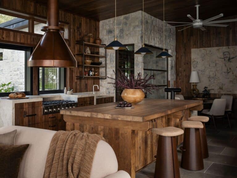 A rustic kitchen features wooden cabinets, a large wooden island with stools, a copper range hood, and pendant lights. Stone walls, open shelves, a cozy sofa, and decorative accents create a warm, inviting atmosphere.