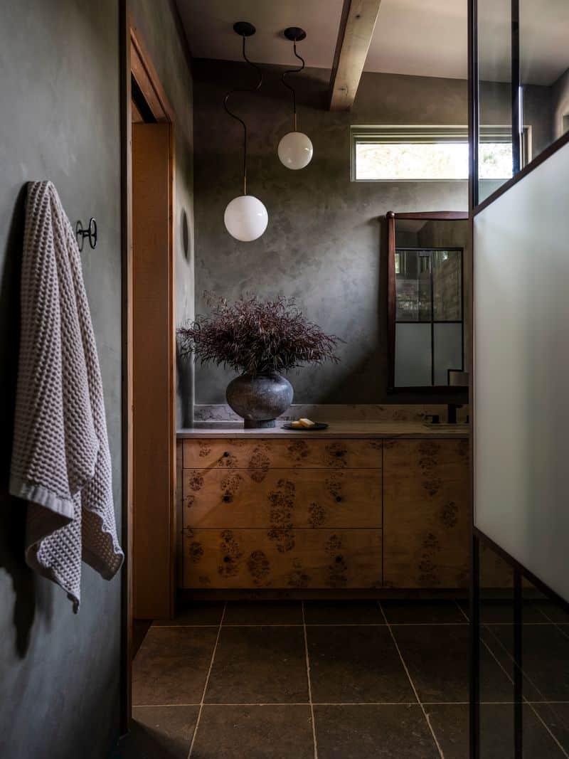 A modern bathroom with dark stone floors, textured gray walls, a wooden vanity with a large vase of foliage, a towel hanging on the wall, and two round pendant lights suspended from the ceiling.