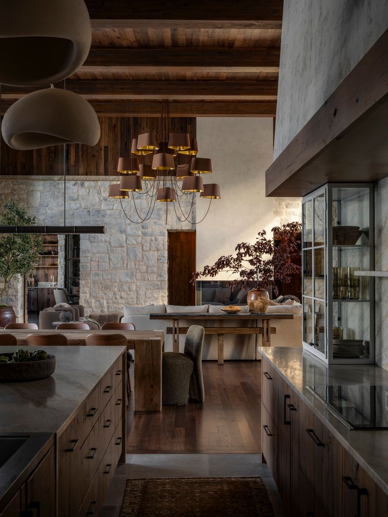 A modern rustic kitchen and dining area with wood beams, stone walls, pendant lights, wooden cabinets, a large chandelier, indoor plants, and a mix of upholstered and wooden furniture. Warm, natural light fills the space.
