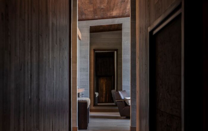 A dimly lit hallway with wooden walls and ceiling leads to a modern living area featuring brown furniture, large windows, and natural light in the distance. Reclaimed Wood Flooring for Sale call Manomin Resawn Timbers