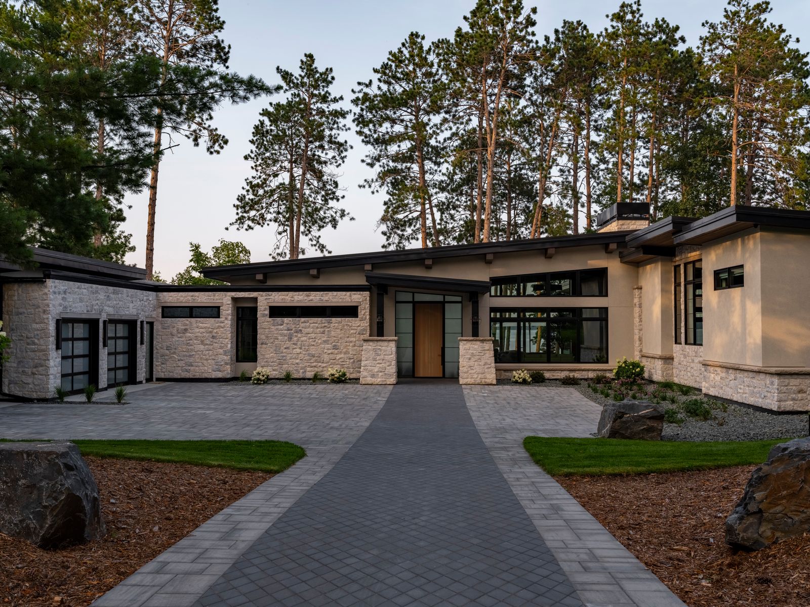 yVW9tLeA Modern house with large windows, stone and stucco exterior, flat roofs, and a landscaped front yard with trees, a paved driveway, and a path leading to the front entrance. Tall pine trees are visible in the background.