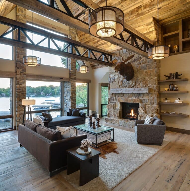 A rustic living room with exposed beams, a stone fireplace, mounted moose head, modern sofas, and large windows with a lake view. Wide-plank-reclaimed-wood-flooring adds character beneath your feet, while warm lighting creates a cozy, inviting atmosphere.