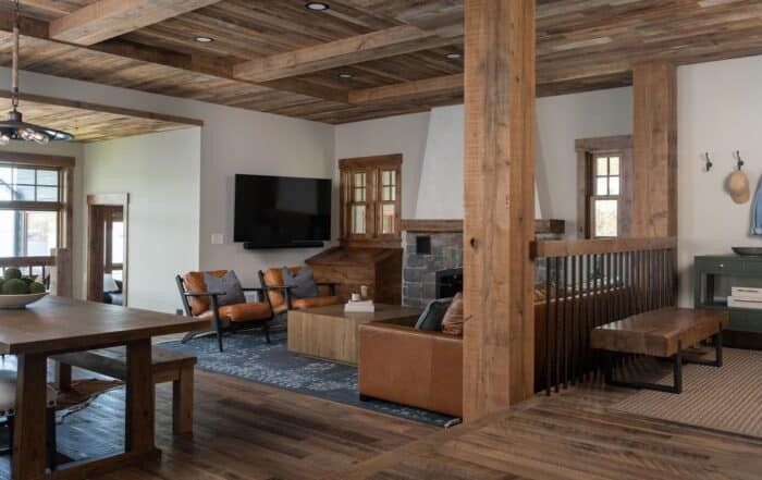 A rustic living room with wooden beams, leather sofas, a wall-mounted TV, and a stone fireplace features wide-plank-reclaimed-wood-flooring that enhances the cozy atmosphere. Large windows and a dining table complement the natural wood tones and neutral colors woven throughout the space.