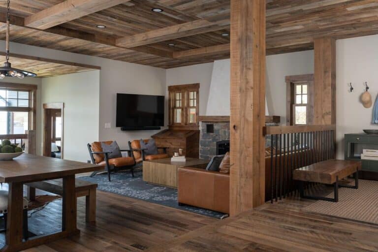 A rustic living room with wooden beams, leather sofas, a wall-mounted TV, and a stone fireplace features wide-plank-reclaimed-wood-flooring that enhances the cozy atmosphere. Large windows and a dining table complement the natural wood tones and neutral colors woven throughout the space.