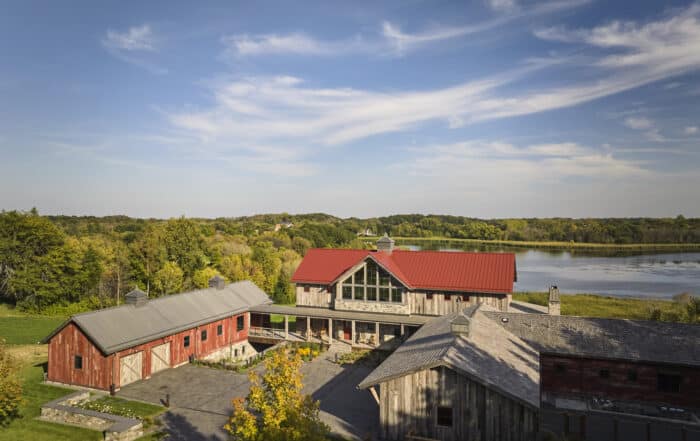 Aerial view of rustic wooden barns with red and gray roofs, surrounded by trees, near a lake under a blue sky with wispy clouds.