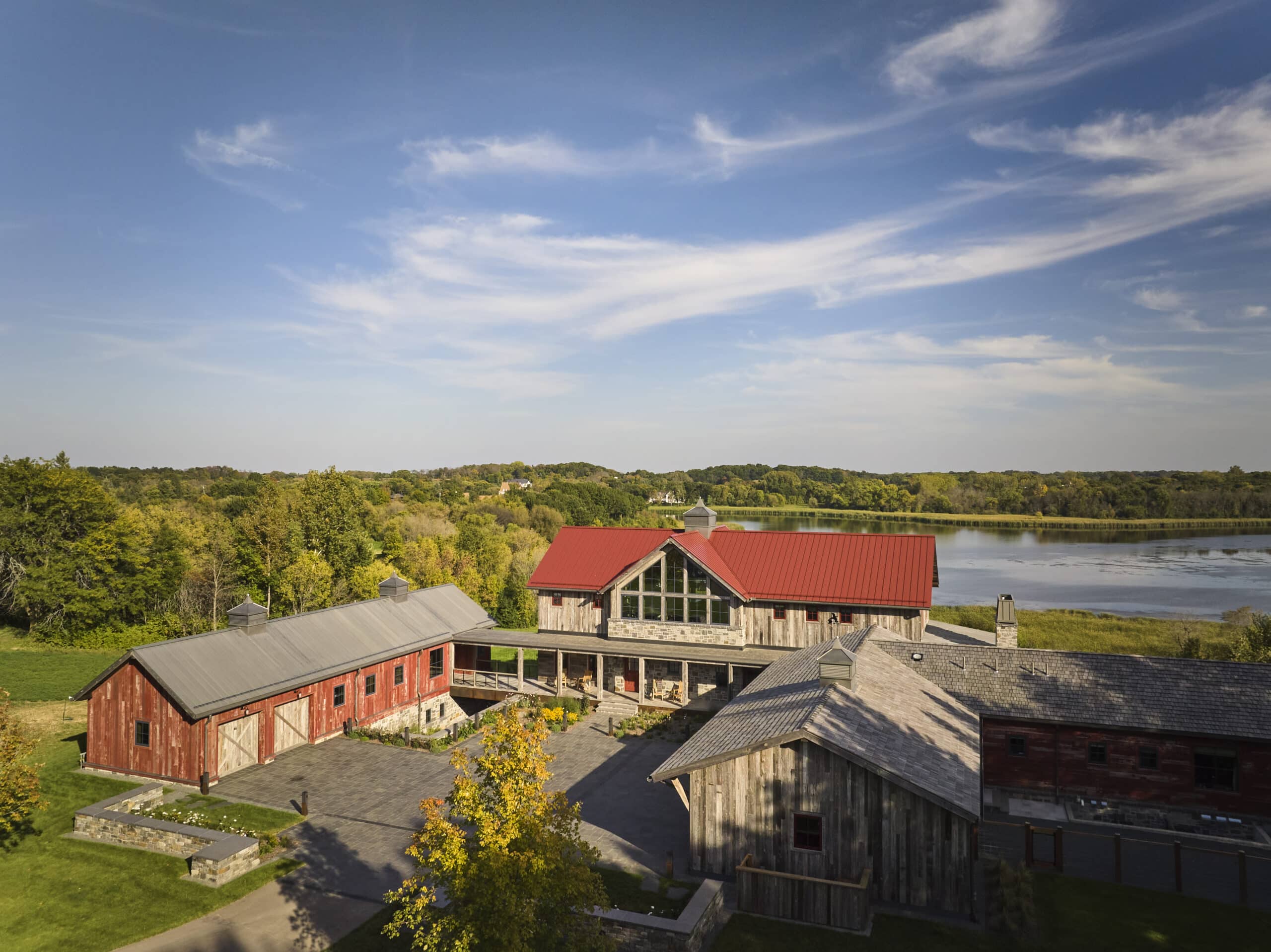 default Aerial view of rustic wooden barns with red and gray roofs, surrounded by trees, near a lake under a blue sky with wispy clouds.