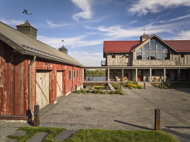 A rustic red barn and a large farmhouse with a red roof stand under a blue sky. The courtyard between them is paved with stone, and there are plants and rocking chairs on the farmhouse porch.