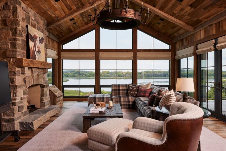 Rustic living room with plaid furniture, stone fireplace, wood ceiling, and large windows overlooking a lake and greenery. Cozy decor includes pillows, candles, a rug, and cow artwork above the fireplace.