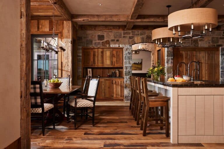 Rustic kitchen and dining area with wooden furniture, stone walls, exposed beams, and two large chandeliers. A kitchen island with stools and fruit, plus a dining table with checkered chairs, are visible. Natural light fills the space.