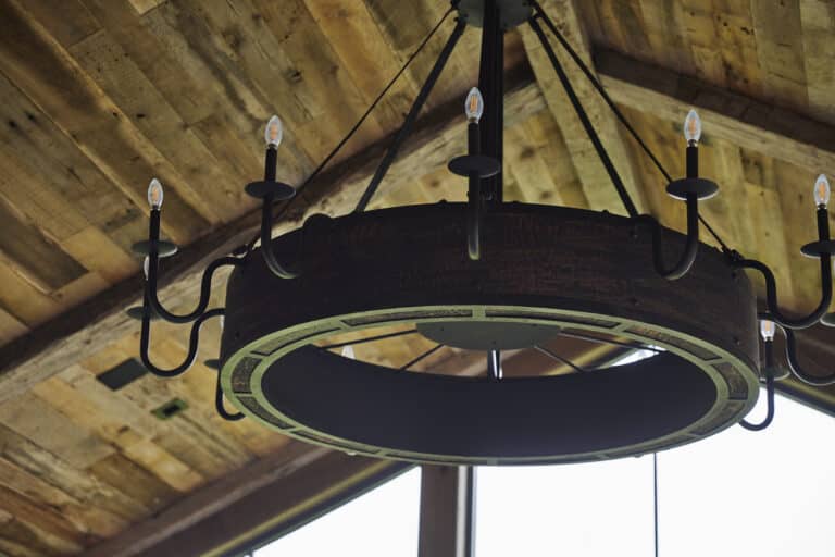 Rustic circular chandelier with candle-shaped LED bulbs, hanging from a wooden ceiling with metal beams visible, creating a cozy cabin-like atmosphere.