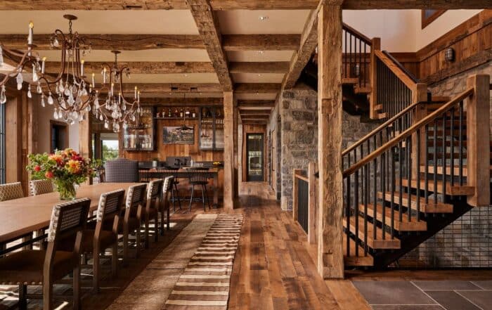 Rustic dining room with a long wooden table, chandelier, wooden beams, stone walls, open staircase, bar area, large windows, and a vase of flowers on the table. Warm lighting and cozy decor create a welcoming atmosphere.