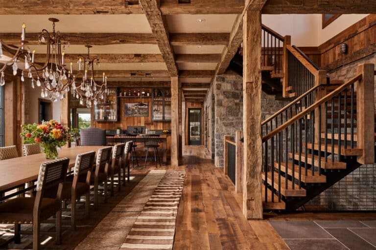 Rustic dining room with a long wooden table, chandelier, wooden beams, stone walls, open staircase, bar area, large windows, and a vase of flowers on the table. Warm lighting and cozy decor create a welcoming atmosphere.