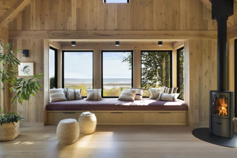 A cozy wooden room with a built-in window seat, cushions, a potted plant, two woven stools, large windows with a scenic outdoor view, and a modern wood stove with a fire burning.