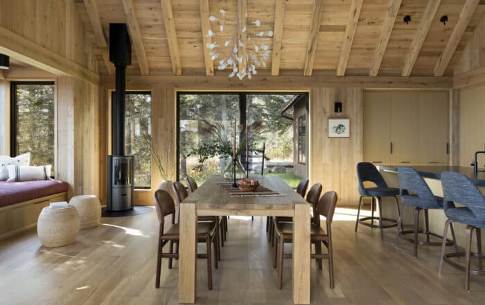 Modern dining room with wood beams, a large wooden table, brown chairs, a black wood stove, blue barstools, large windows, natural light, and a nature view outside. Decor includes plants, a chandelier, and woven baskets.