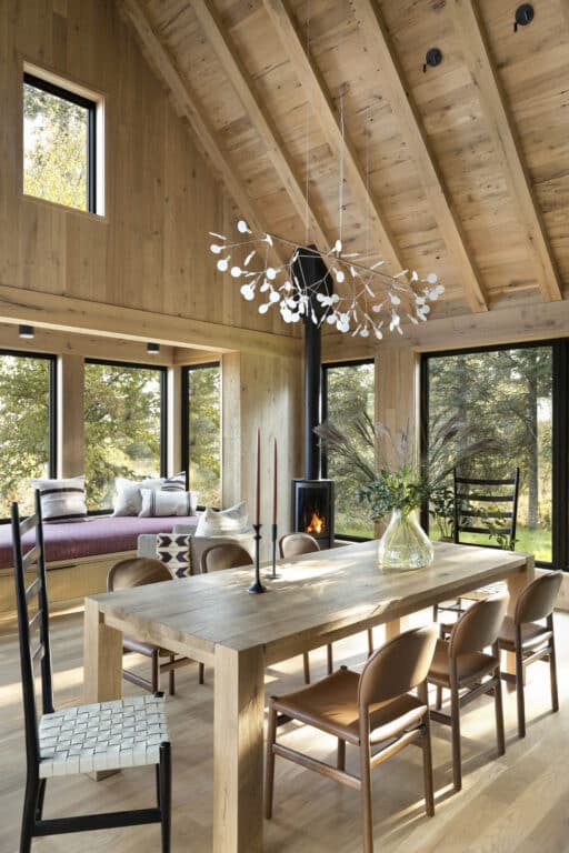 A modern dining room with a wooden table, six brown chairs, a black chair, pendant light, wood stove, large windows, and a cozy window seat with cushions, all set in a high-ceilinged wooden interior.