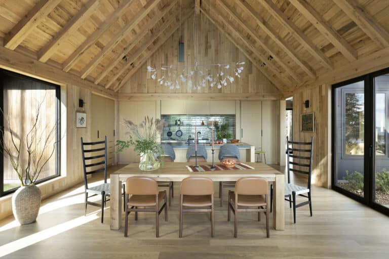 A wooden dining room with a vaulted ceiling, a wooden table set for six, two tall black chairs at each end, modern kitchen in the background, large windows, and natural light filling the space.