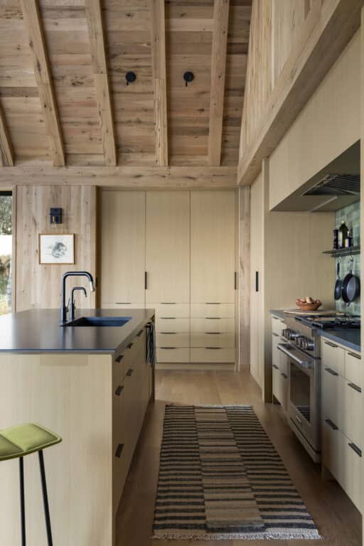 Modern kitchen with light wood cabinets, black fixtures, a striped rug, island seating, exposed wooden beams on the ceiling, and open shelving with dishes. Natural light enters from a window on the left.