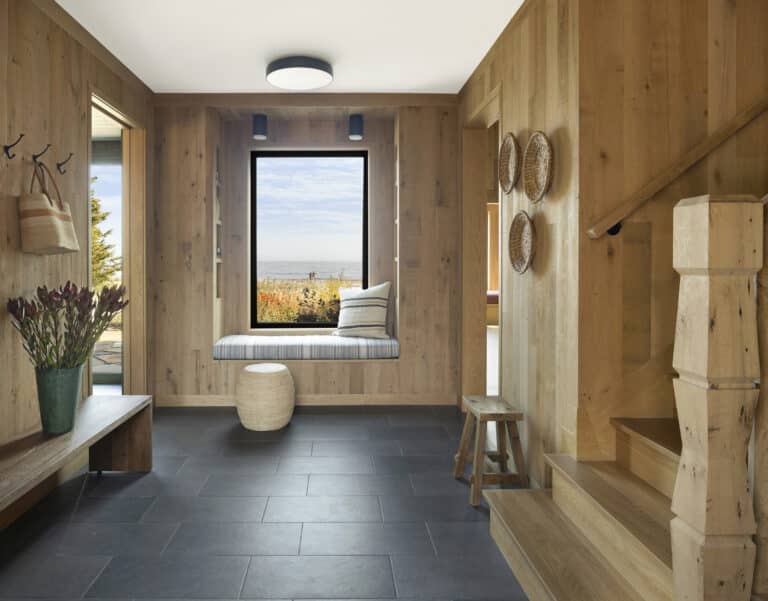 A modern entryway with wood-paneled walls, a window seat, a small stool, baskets on the wall, a bench with a vase of flowers, and stairs leading up, overlooking a scenic outdoor view.