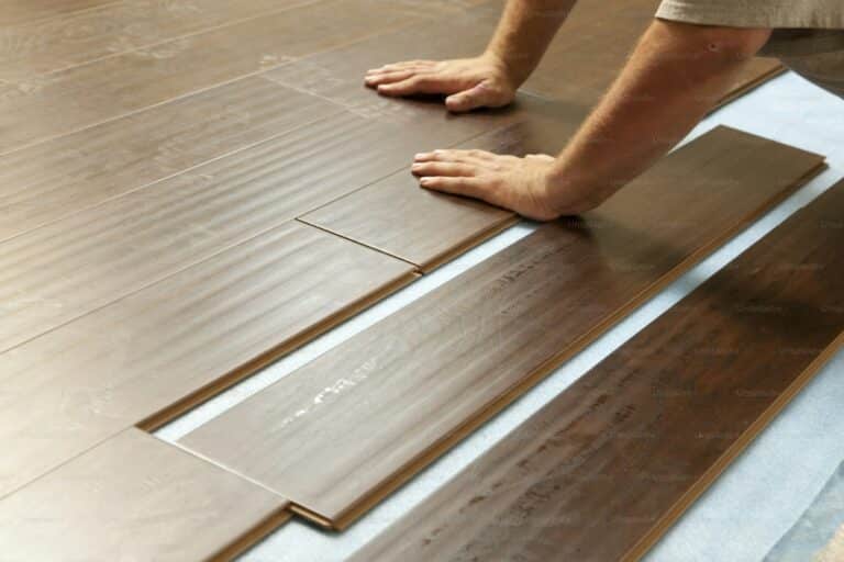 Close-up of a person installing wooden laminate flooring, using their hands to position and press the planks together over a blue underlayment.